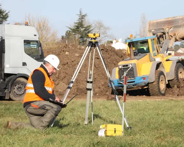Geodeta w kamizelce odblaskowej nadzoruje odwierty geologiczne pod budowę domu. W tle koparka i ciężarówka.