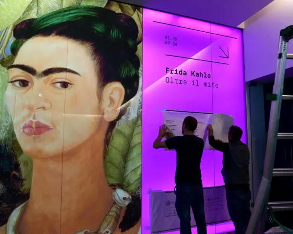 Un portrait de Frida Kahlo, avec deux hommes installant une affiche pour une exposition sur son histoire.