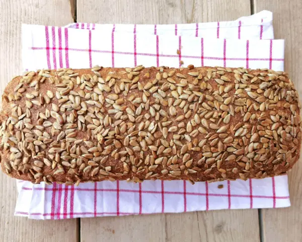Ein rustikales Brot, bedeckt mit vielen Sonnenblumenkernen, liegt auf einem karierten Küchentuch.