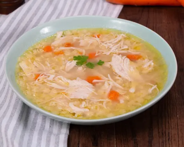 Un confortevole brodo di pollo con pezzi di pollo sfilacciato, carote e un rametto di prezzemolo, servito in una ciotola azzurra.