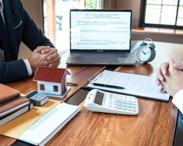 Réunion pour discuter de la faute du mandataire de gestion locative : maison modèle, calculatrice, documents et ordinateur portable sur un bureau en bois.
