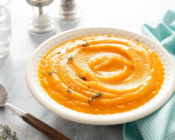 Un bol de velouté de purée de butternut, garni de thym frais, prêt à être dégusté avec une cuillère en argent.