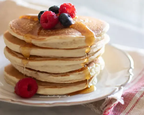 Pancakes senza uova dorati, impilati e inzuppati di sciroppo, guarniti con lamponi e mirtilli freschi.
