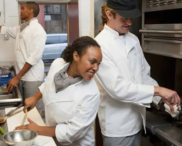 La brigada de cocina y sus funciones: chefs preparan alimentos, uno corta vegetales, otro cocina en sartén y otro lava utensilios.