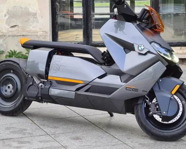 Szary skuter elektryczny BMW CE 04 z pomarańczowymi akcentami stoi na bruku przed budynkiem z dużymi oknami.