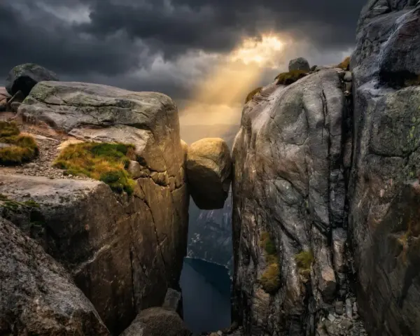 Enorm sten balanserar mellan klippor i Norge. Dramatiskt ljus bryter igenom molnen över en djup fjord.
