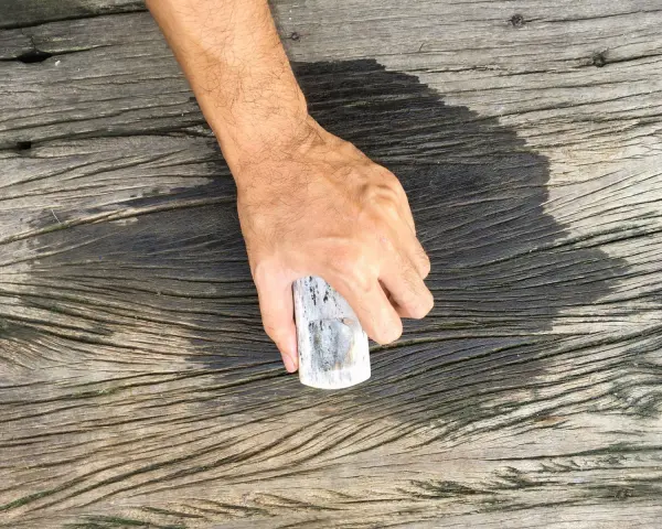 En hand håller en borste för att skura altanen. Träet är väderbitet och har en mörk, fuktig yta där borsten nyss varit.