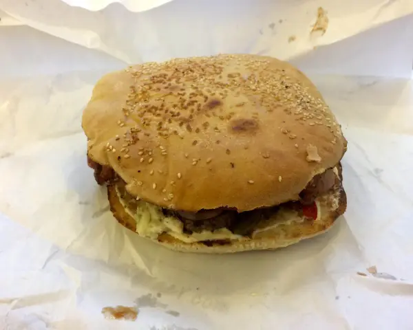 Burger z sezamem na białym papierze, widoczne składniki: mięso, sos, warzywa.