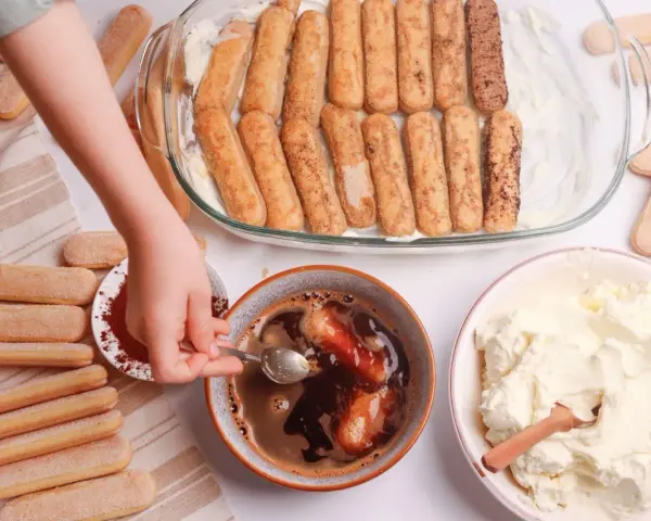 Dziecko zanurza biszkopty w kawie, przygotowując warstwy do tiramisu. Obok krem i kakao.