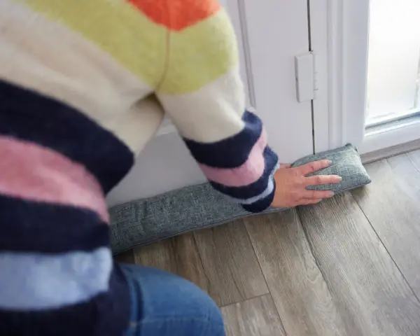 Ocieplanie drzwi wejściowych: osoba w kolorowym swetrze układa podkładkę izolacyjną pod drzwiami, by zatrzymać przeciągi.
