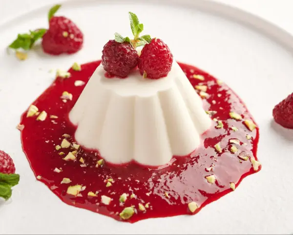 Panna Cotta przepis: kremowy deser z malinami, pistacjami i miętą na białym talerzu.