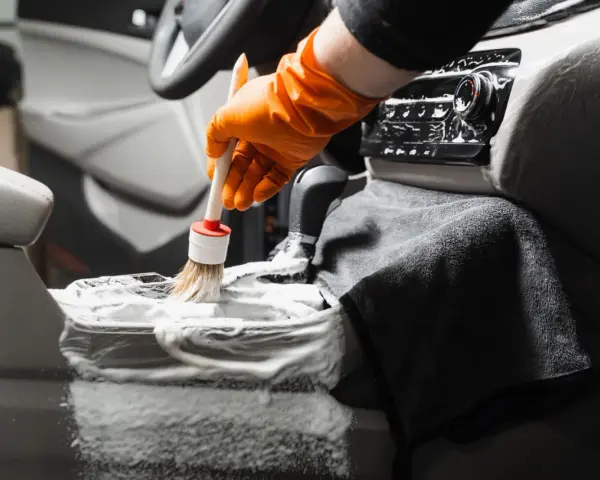 En hand med orange handske använder en **detail brush** för att rengöra bilens mittkonsol med skum.