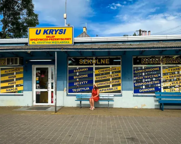 Kobieta w czerwonej sukience siedzi na ławce przed sklepem "U Krysi", na którym widnieją napisy "Szkolne" i "Narzędzia".