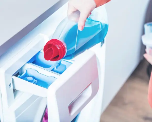 Eine Hand gießt flüssiges Waschmittel in das Fach einer Waschmaschine. So benutzt man eine Waschmaschine richtig.