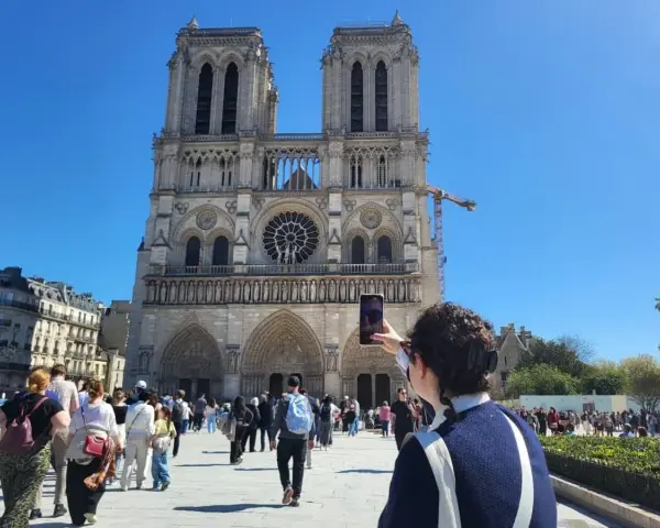 Una persona toma una foto de la fachada de Notre Dame. Mucha gente pasea por la plaza, disfrutando de su visita a Notre Dame.