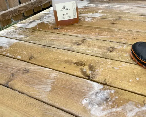 En flaska linoljesåpa står på en altan som håller på att rengöras. En svart stövel med orange kant syns i nedre högra hörnet.