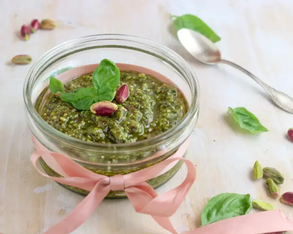 Un vasetto di pesto di pistacchi, decorato con un nastro rosa e guarnito con foglie di basilico e pistacchi interi, pronto per essere gustato.