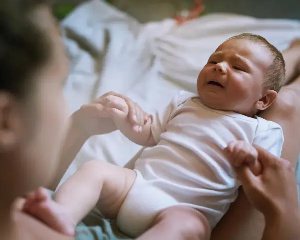 Płaczliwe niemowlę w białym body, trzymające mamę za rękę. Co robić, gdy pojawia się histeria u niemowlaka?