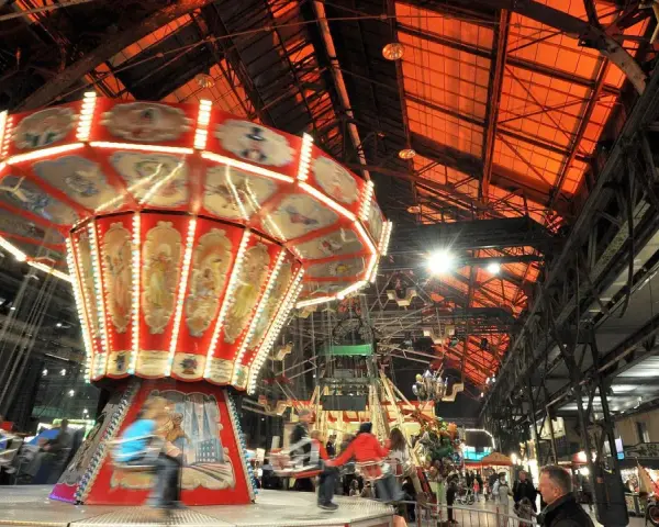 Drehende Kettenkarussells und bunte Lichter auf einem historischen Jahrmarkt in einer alten Industriehalle.