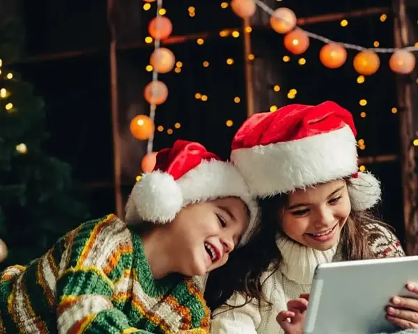 Dos niños con gorros de Santa Claus disfrutan de la tablet, felices cuando dan vacaciones de Navidad, rodeados de luces festivas y un árbol.