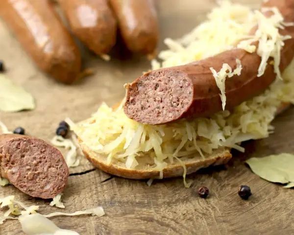 Lungenwurst zubereiten: Eine Scheibe Brot mit Sauerkraut und Lungenwurst belegt, daneben weitere Würste, Gewürze und Lorbeerblätter auf Holzuntergrund.