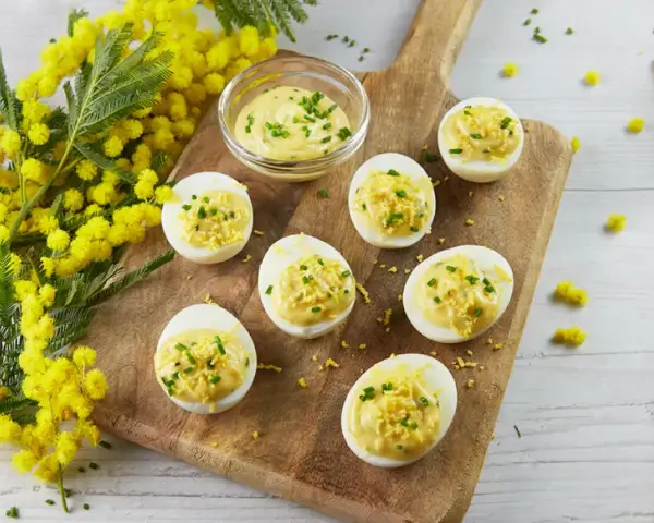 Œufs mimosa garnis d'une sauce crémeuse et de ciboulette, présentés sur une planche en bois avec des fleurs de mimosa. Une recette printanière.