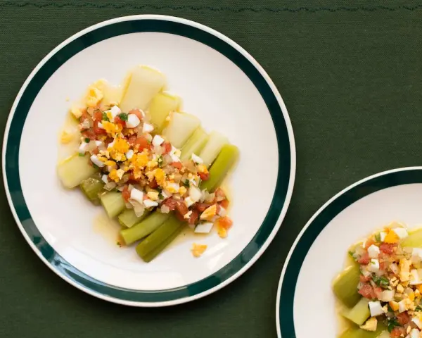 Puerros a la vinagreta servidos en platos blancos con borde verde, cubiertos con una mezcla de tomate picado, huevo duro y perejil.