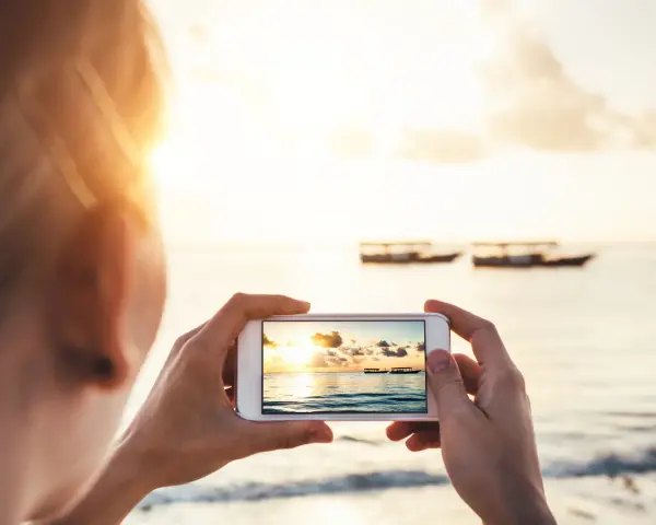 Une personne prend une photo d'un coucher de soleil sur la mer avec des bateaux au loin. Une belle photo à faire à la plage.