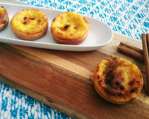 Vier goldbraune, karamellisierte Pastéis de Nata auf einem weißen Teller, daneben Zimtstangen auf einem Holzbrett.