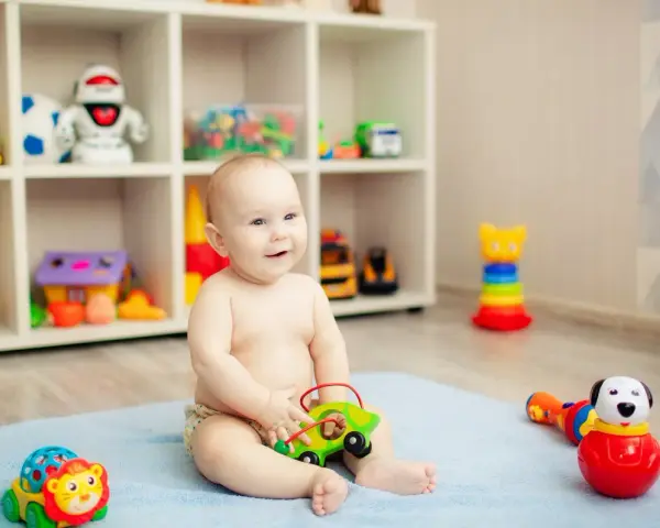 Uśmiechnięte, 8-miesięczne dziecko nie siedzi, bawi się zielonym autkiem na niebieskim kocyku. W tle regał z zabawkami.