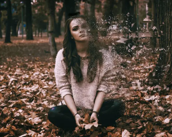 Kobieta w swetrze siedzi na jesiennych liściach, jej ciało rozpada się na fragmenty.