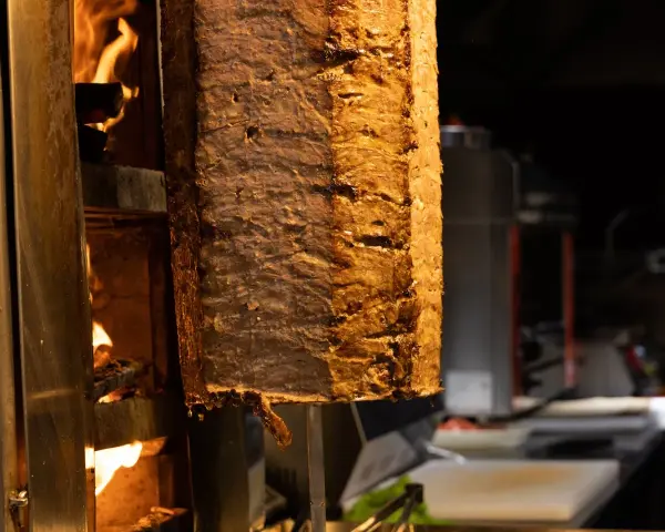 Duży pionowy obrotowy grill z mięsem, obok płomienie ognia.