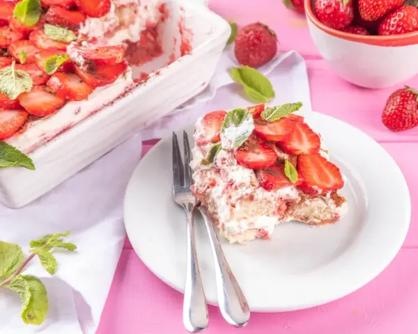 Tiramisu klasyczne kontra tiramisu truskawkowe. Porcja deseru z truskawkami i miętą na talerzu, obok naczynie z deserem i miseczka owoców.