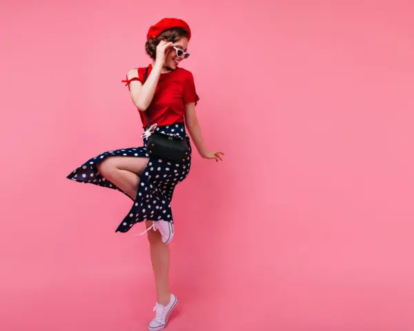 Kobieta w stylu pin-up w czerwonym berecie i sukience w grochy skacze na różowym tle.