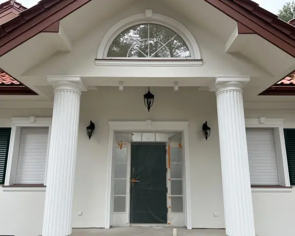Elegancki pilaster zdobi fasadę domu, wspierając dach nad wejściem. Obok okna i drzwi, tworzy klasyczną kompozycję.