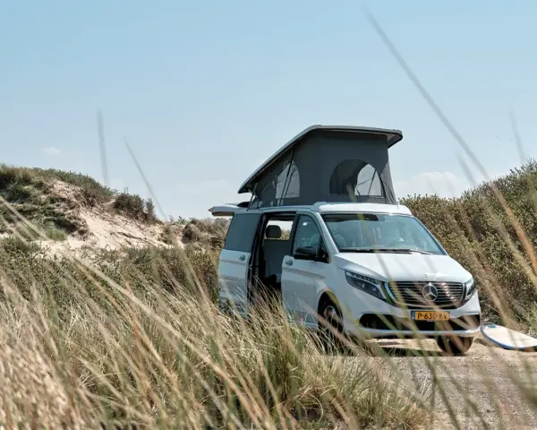 Vit el campervan med uppfällt tak står parkerad vid en sanddyn. En surfbräda ligger bredvid.