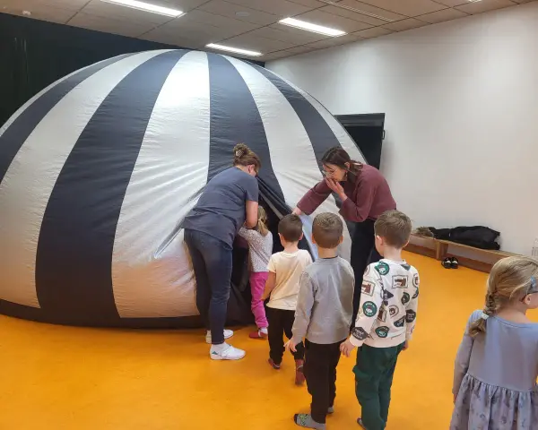 Dzieci i opiekunki wchodzą do dmuchanej kopuły, która przypomina planetarium. To miejsce, gdzie można oglądać filmy z przesłaniem, odkrywając tajemnice wszechświata.