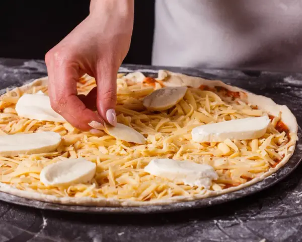 Dłoń układa kawałki sera na pizzy, która jest już posypana startym żółtym serem.