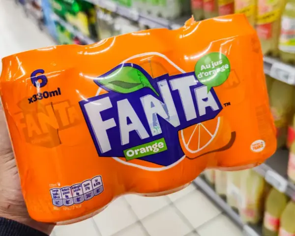 Opakowanie 6 puszek Fanty Orange w ręce, na tle sklepowych półek z napojami.