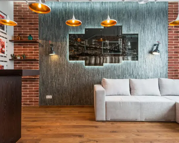 Nowoczesny salon w stylu loft: szara kanapa, ceglana ściana, panel z nocnym widokiem miasta i złote lampy.