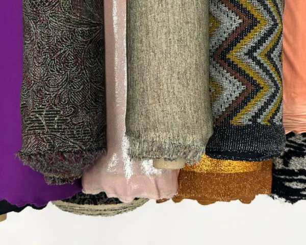 Rouleaux de tissus aux textures variées : noir uni, violet, motifs complexes, rose pâle, beige chiné, zigzags multicolores, pêche, et noir et blanc. Une matière mesh se distingue par sa finesse.
