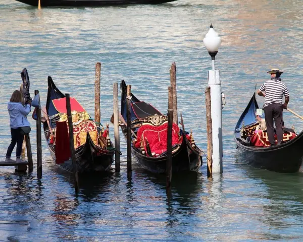 Gondole veneziane ornate, una turista scatta foto. La differenza tra gondola e sandalo è chiara: una naviga, l'altro si indossa.