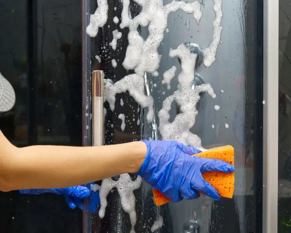 Main bleue, éponge orange pour nettoyer paroi douche. Mousse et bulles sur la paroi vitrée.