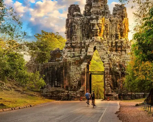 Two cyclists approach a massive stone gateway, one of the ancient Cambodia temples, bathed in golden sunlight.