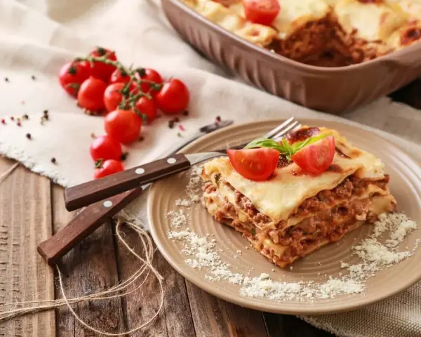 Pyszna, wielowarstwowa lasagne z mięsem mielonym, polana sosem beszamelowym i posypana parmezanem. Na talerzu ozdobiona pomidorkami koktajlowymi i bazylią.