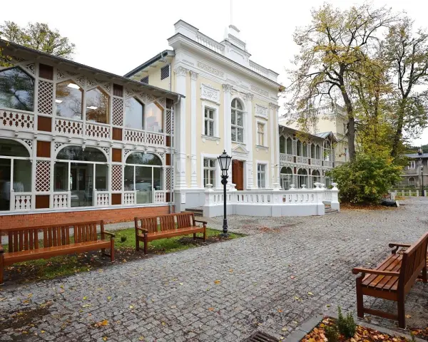 Elegancki budynek z ażurowymi werandami, przypominający dawny **Hevelianum dom zdrojowy**. Brukowany dziedziniec z ławkami i jesiennymi liśćmi.