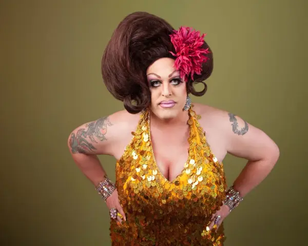 Osoba w złotej, cekinowej sukience i peruce, z kwiatem we włosach. To przykład tego, co to jest drag queen – artystyczna forma ekspresji.