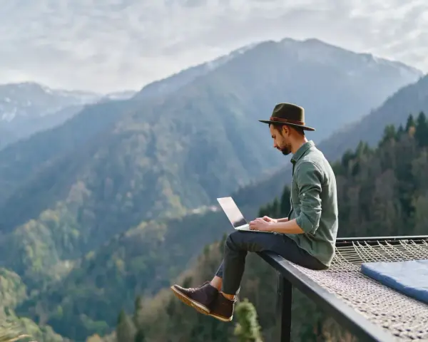 Un homme en chapeau travaille sur son ordinateur portable, profitant du télétravail à l'étranger avec une vue imprenable sur les montagnes.