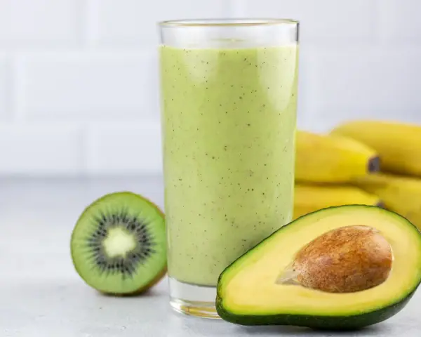 Zielony koktajl z awokado, kiwi i bananów w szklance, obok połówka awokado i kiwi.