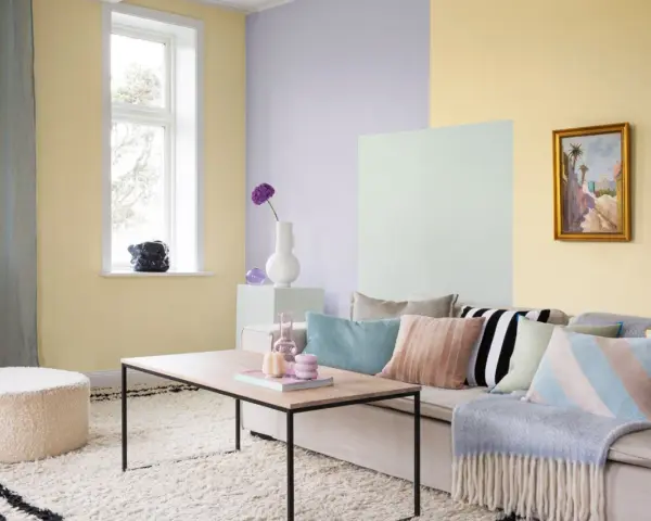 Ett vardagsrum med pastell inredning. Ljusgula, ljuslila och ljusgröna väggar skapar en mjuk atmosfär. En beigefärgad soffa är fylld med kuddar i olika pastellfärger och mönster.